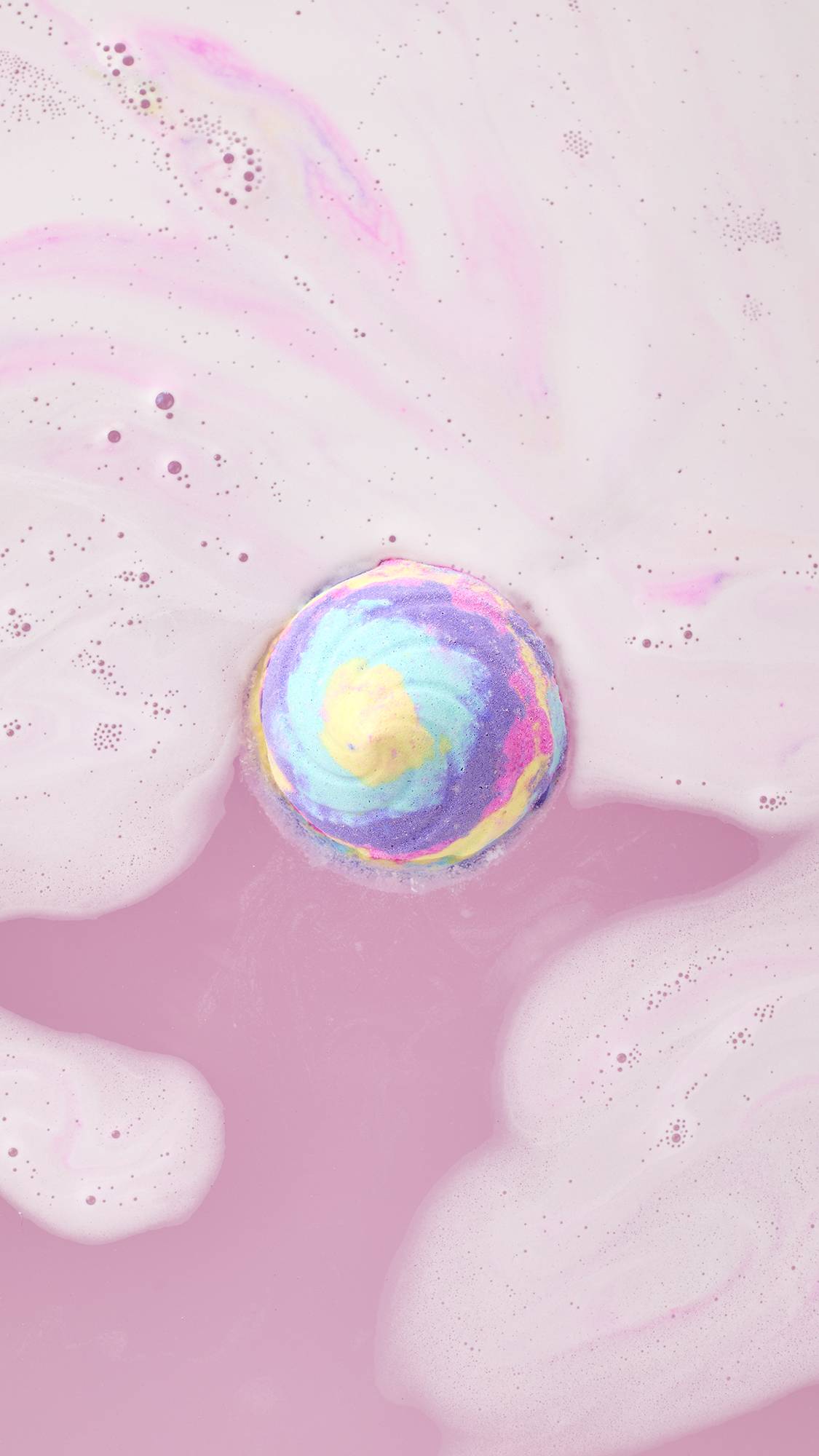 Top-down view of the Unicorn Poop Bath Bomb floating in a pool of milky pink bathwater, releasing soft swirls of pastel yellow, blue, and purple foam.