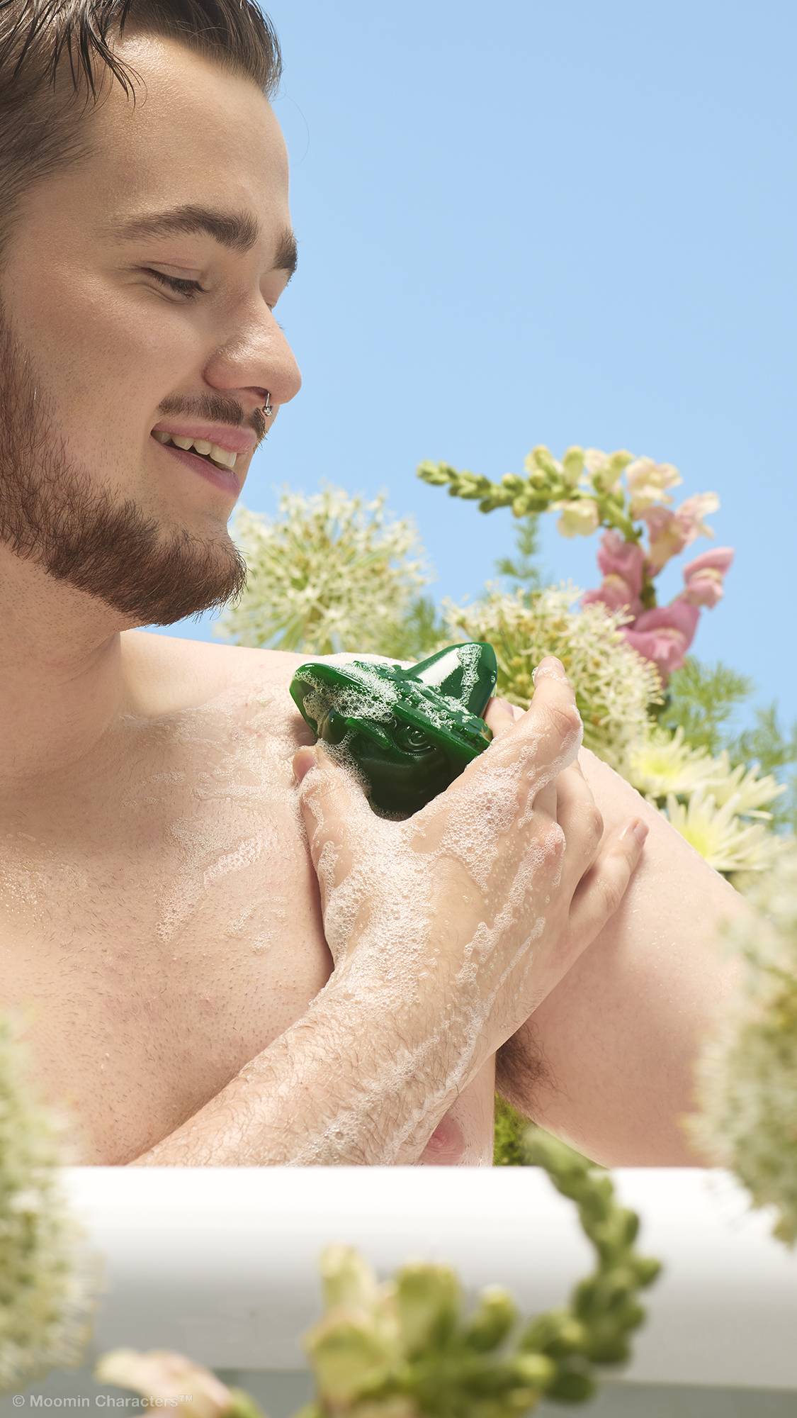 A close-up shot of a smiling person with a beard in a white bathtub, using a green Snufkin-shaped shower gummy to lather their shoulder. White and pink wildflowers are visible in the background against a clear blue sky.