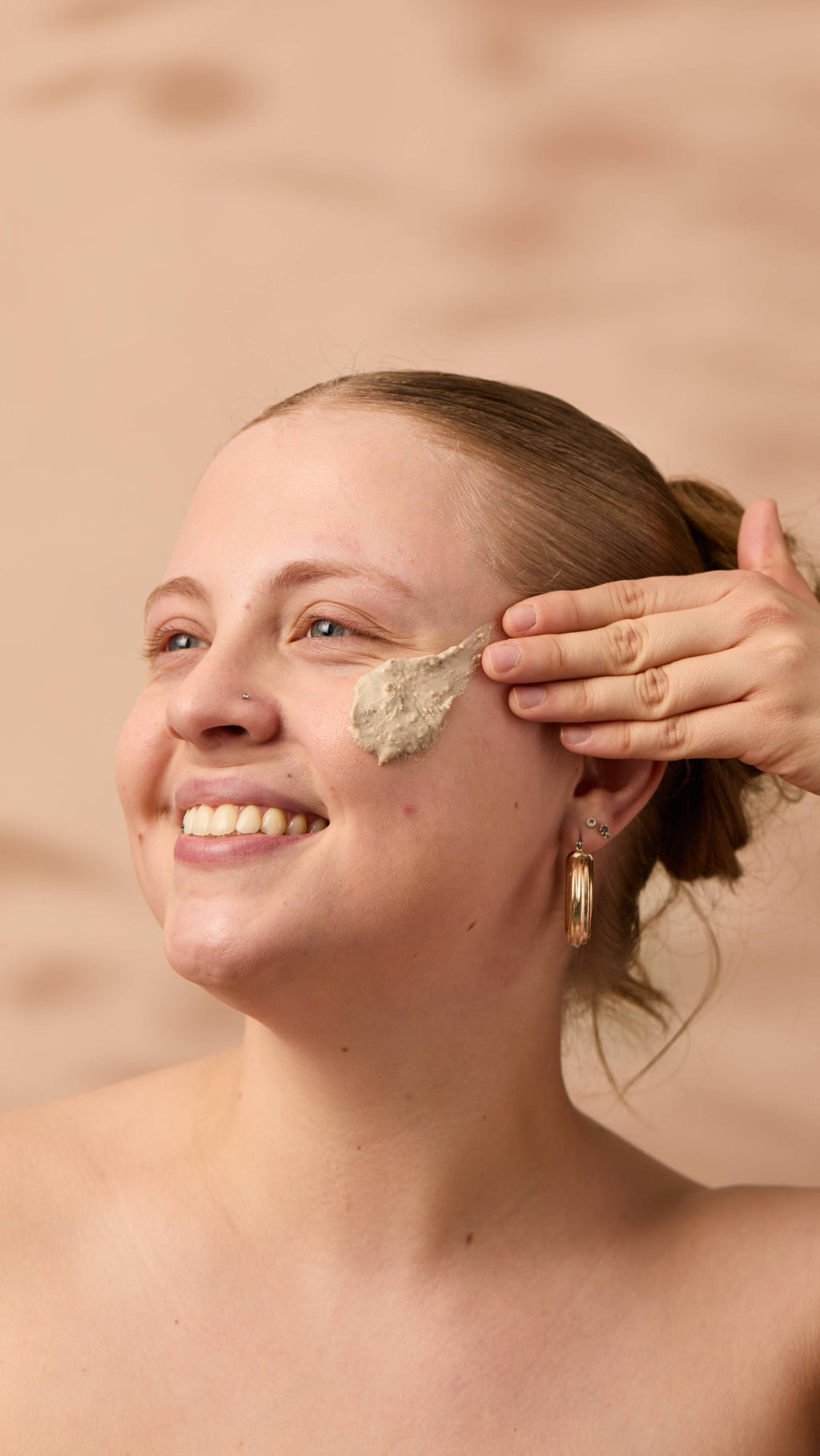 An image of the model's face on a warm, beige background. Their copper hair is tied back, and they have applied the textured Bring Us Some Figgy Pudding face mask onto their cheek as they smile.
