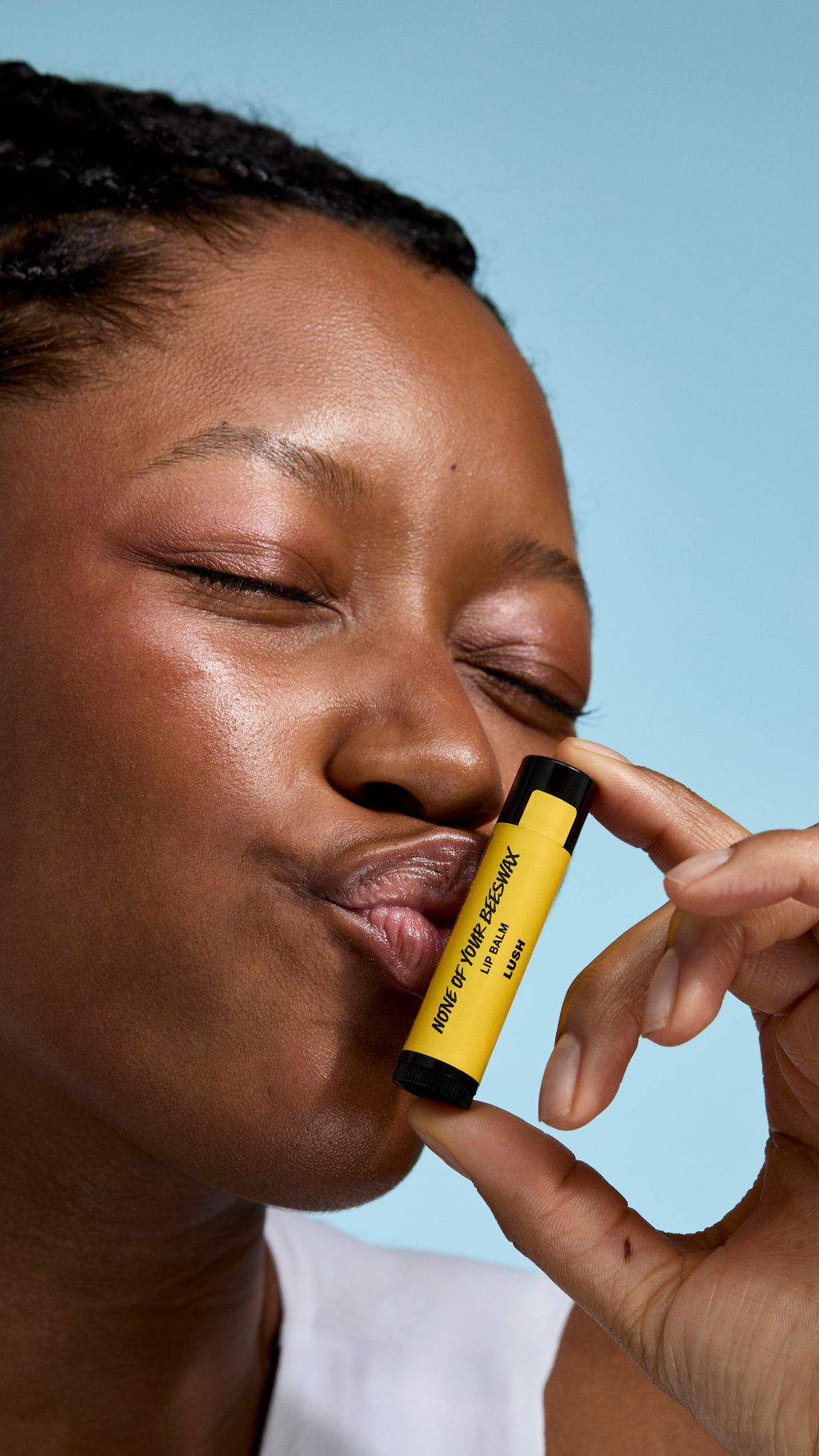 A close-up of a model with deep-toned skin and braided hair holding a yellow 'None of Your Beeswax' lip balm. They have their eyes closed and are making a playful kissing face, with their lips touching the tube against a soft blue background.