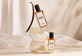 The image shows a background of layered, cream, satin fabric. In the centre is the focus of precisely balanced, white stacked stones. There is a dried vanilla pot standing on top and the two sizes of perfume leaning against the stones.