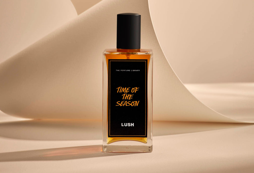 The image shows the Time Of The Season glass perfume bottle. It is sitting on a soft, beige background with curved, cream paper creating soft, gliding shapes behind the product.